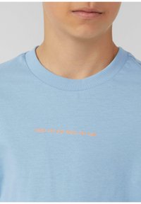 Lichtblauwe katoenen T-shirt met een ronde hals en korte mouwen, met oranje tekst: "Jaag de zon achterna, geniet van het plezier," gedrukt aan de voorkant.