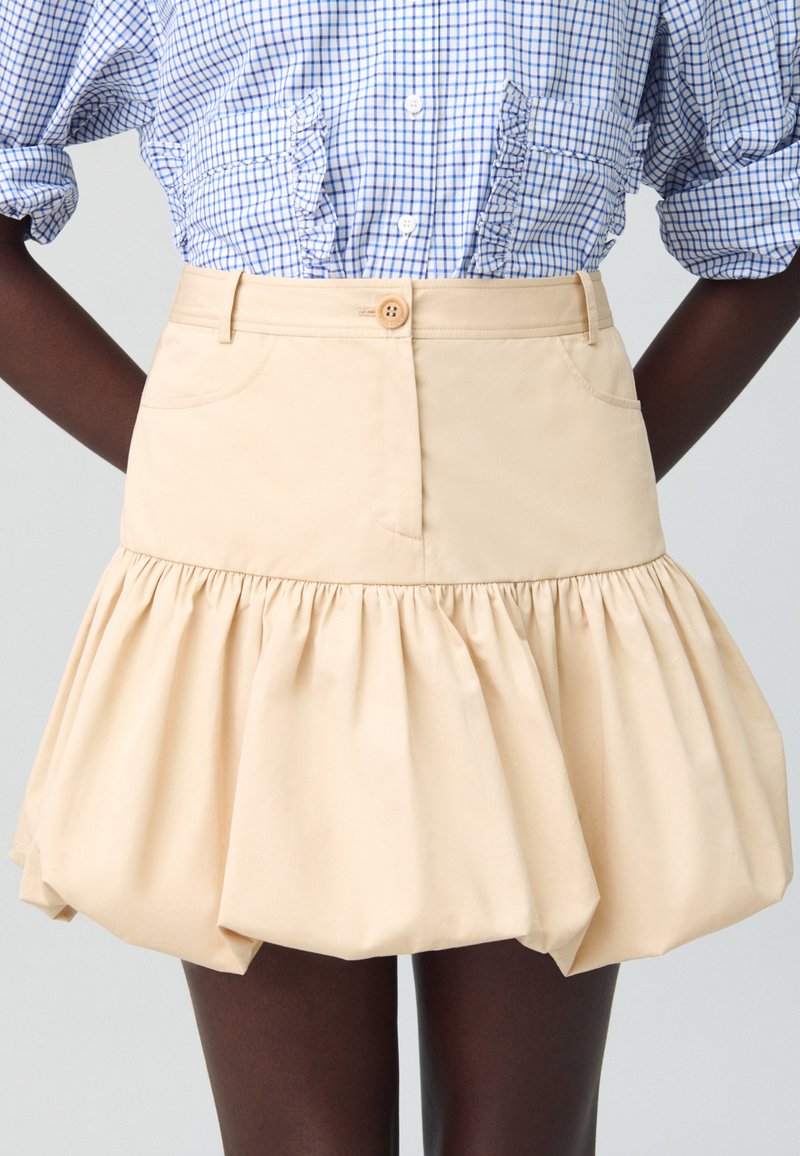 Jupe en coton beige avec un ourlet en volants évasé, dotée d'une fermeture à boutons sur le devant et de poches latérales. Associée à une chemise à motif �à carreaux bleus.