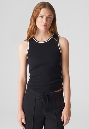 Schwarzes, geripptes Tank-Top mit weißen Akzenten am Halsausschnitt und den Armausschnitten, taillierte Passform. Kombiniert mit schwarzen Sweatpants mit Kordelzug, glatte Textur.
