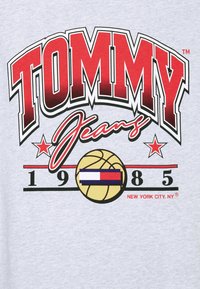 Punainen ja musta "TOMMY" -teksti kursiivilla "Jeans" -sanan yllä, jonka päällä on koripallo, jossa on punainen, valkoinen ja sininen lippu, tähtiä ja "1985 New York City, NY."