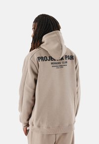 Sudadera beige con un gran estampado gráfico negro en la parte posterior, hecha de una tela suave con un corte relajado y capucha ajustable.