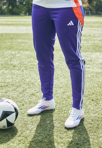 Purple athletic pants with white and blue stripes, paired with white soccer cleats featuring red and blue accents, next to a black and white soccer ball.