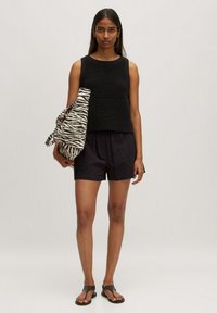 Zwarte gebreide tanktop gecombineerd met zwarte gestructureerde shorts. Model houdt een tas met zebra-print vast. Zwarte sandalen maken de outfit compleet.