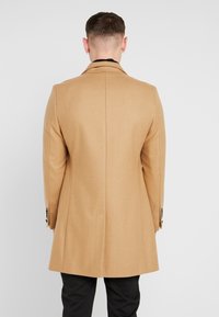 Wollmantel in Camel mit geradem Schnitt, featuring einen Reverskragen und besetzten Bündchen, von hinten vor einem schlichten Hintergrund betrachtet.