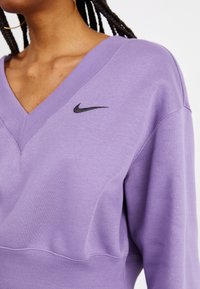 Person som bär en lavendel V-ringad sweatshirt med en svart Nike-swoosh-logotyp och guldörhängen, visar flätat hår och nacke.
