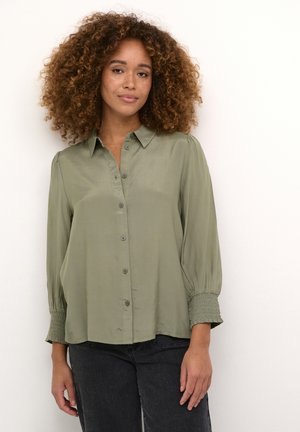 Groene blouse met knopen, een puntige kraag, lange mouwen met gerimpelde manchetten en een gladde, lichte stofstructuur.