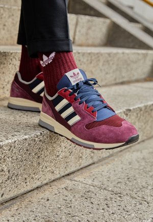 Osoba nosząca bordowe sneakersy Adidas z granatowymi i białymi paskami, w połączeniu z pasującymi bordowymi skarpetkami Adidas, stawiająca krok na betonowych schodach.