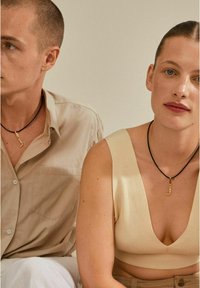 Collier en cordon noir avec un pendentif en or, porté par deux mannequins. La femme porte un haut crème à côtes ; l'homme est en chemise beige à boutons.