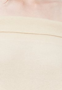 Top de malha canelada bege com design fora dos ombros, apresentando uma textura suave e uma costura horizontal ao longo do decote.