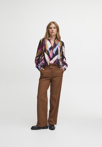 Blusa multicolore con motivi geometrici; pantaloni neri e marroni; scarpe lucide nere. La blusa presenta maniche lunghe e un colletto.