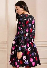 Črna obleka s cvetličnim vzorcem v roza, vijolični in rdeči barvi, z pikami na rokavih, z nabranim pasom in razširjenim robom. Gladka tekstura tkanine.