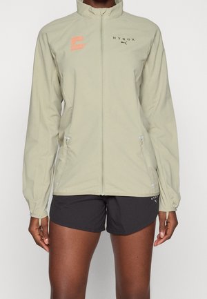 Personne portant une veste zippée beige clair avec les logos « HYROX » et « World Series of Fitness Racing » ainsi qu'un short de sport noir.