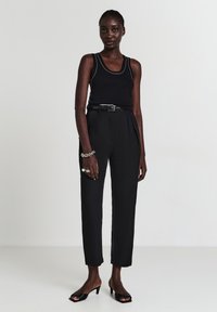 Donna che indossa un top senza maniche nero, pantaloni neri a vita alta con cintura, bracciale e anelli argento, in piedi su tacchi aperti neri su sfondo bianco.