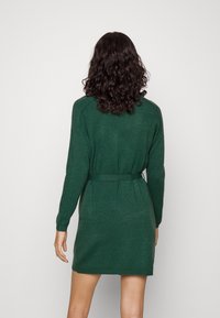 Robe verte tricotée avec une taille ceinturée, des manches longues et des poignets côtelés. Texture lisse et tissu léger, conçue pour une silhouette ajustée.