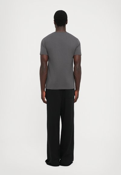 Man die met zijn rug naar de camera staat, gekleed in een aansluitend grijs T-shirt met korte mouwen en losse zwarte broek, staand tegen een egale witte achtergrond.