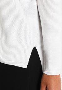 Detalle de suéter blanco de punto que muestra textura acanalada, abertura lateral y mangas largas, combinado con un fondo negro.