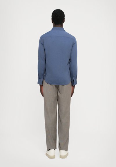 Langdurige, blauwe overhemden met lange mouwen en een rechte zoom, gedragen met lichtgrijze geruite broek en witte schoenen, gezien vanuit de achterkant.