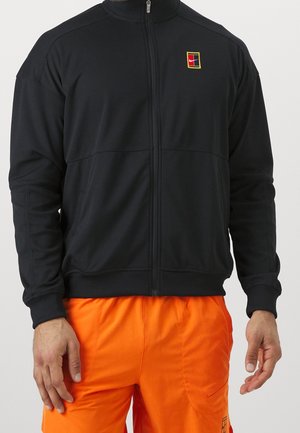 Man draagt een zwart jack met rits en een klein kleurrijk logo op de borst en feloranje broek tegen een effen achtergrond.