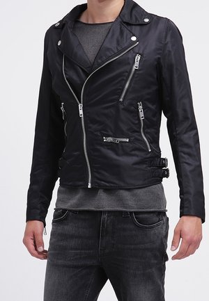 Black asymmetrical zip-up biker jacket with multiple silver zipper pockets, worn over a gray sweater and paired with dark gray jeans.