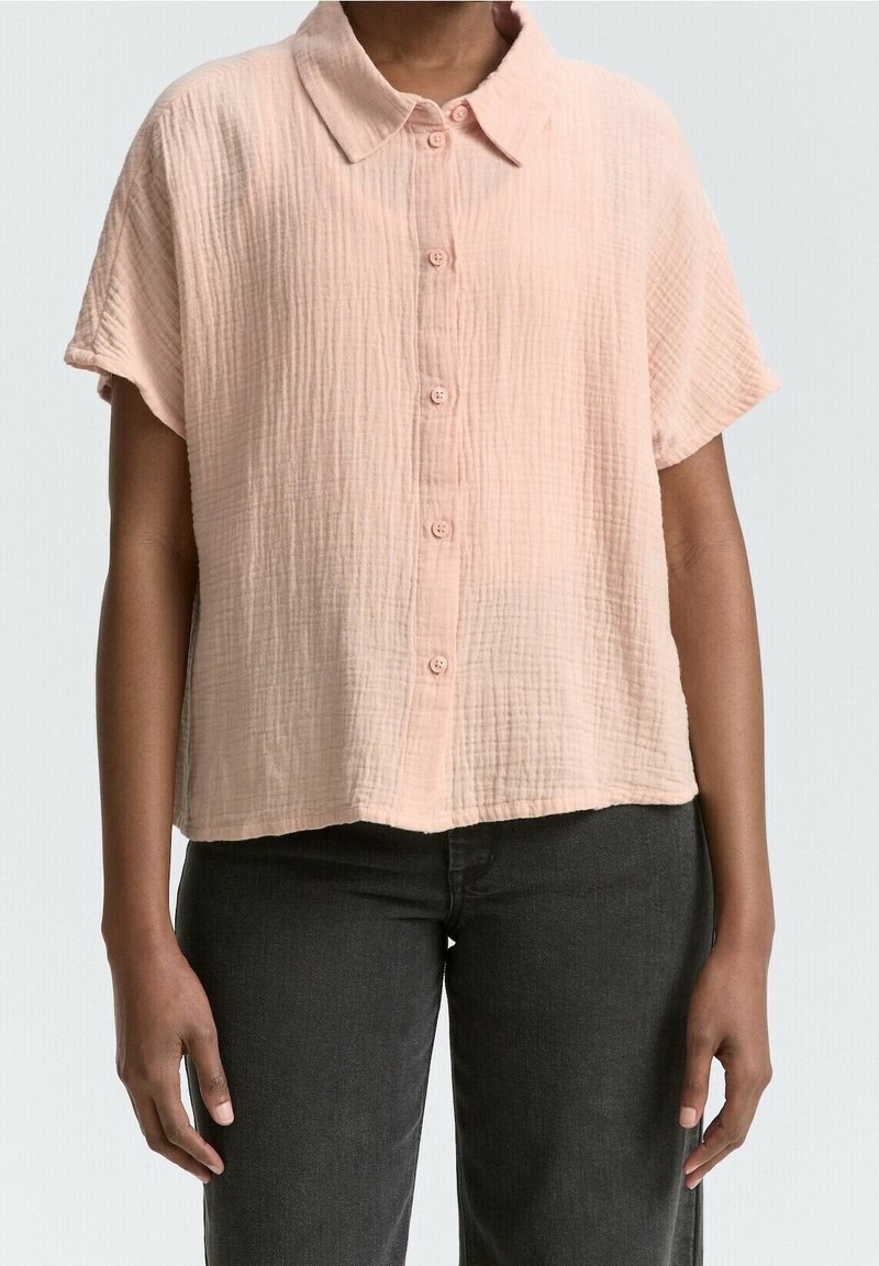 Femme portant une chemise boutonnée à manches courtes, rose pâle texturée avec col, et un pantalon noir sur un fond uni.