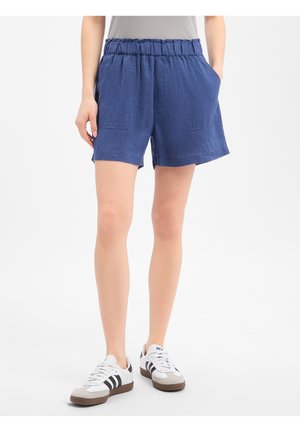 Personne portant un short bleu à taille élastique avec des poches devant et des baskets blanches avec des rayures noires sur fond blanc.