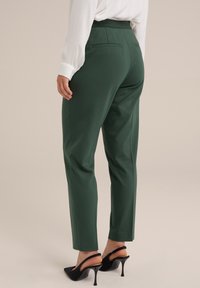 Groene op maat gemaakte broeken met een gladde textuur, taps toelopend been en twee achterzakken. Draagt een witte blouse met zwarte hoge hakken.