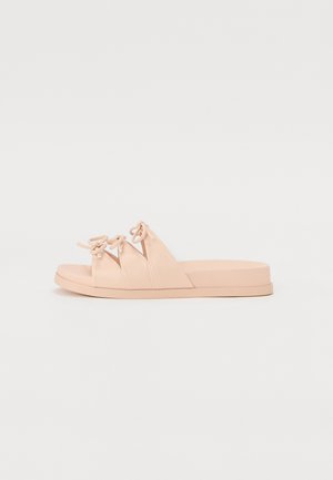 Sandales roses avec trois brides superposées présentant des détails de nœud, une semelle plate et une texture lisse. Design minimaliste, construction légère.