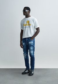 Άσπρο βαμβακερό T-shirt με κίτρινο σχέδιο χαμογελαστής φατσούλας, σκισμένο μπλε τζιν, μαύρα παπούτσια με λευκές ρίγες και μεγάλα γυαλιά ηλίου.