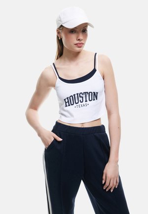 Biały krótki top na ramiączkach z napisem "HOUSTON TEXAS" w granatowym kolorze, zestawiony z granatowymi spodniami z białymi paskami po bokach. Modelka nosi białą czapkę.