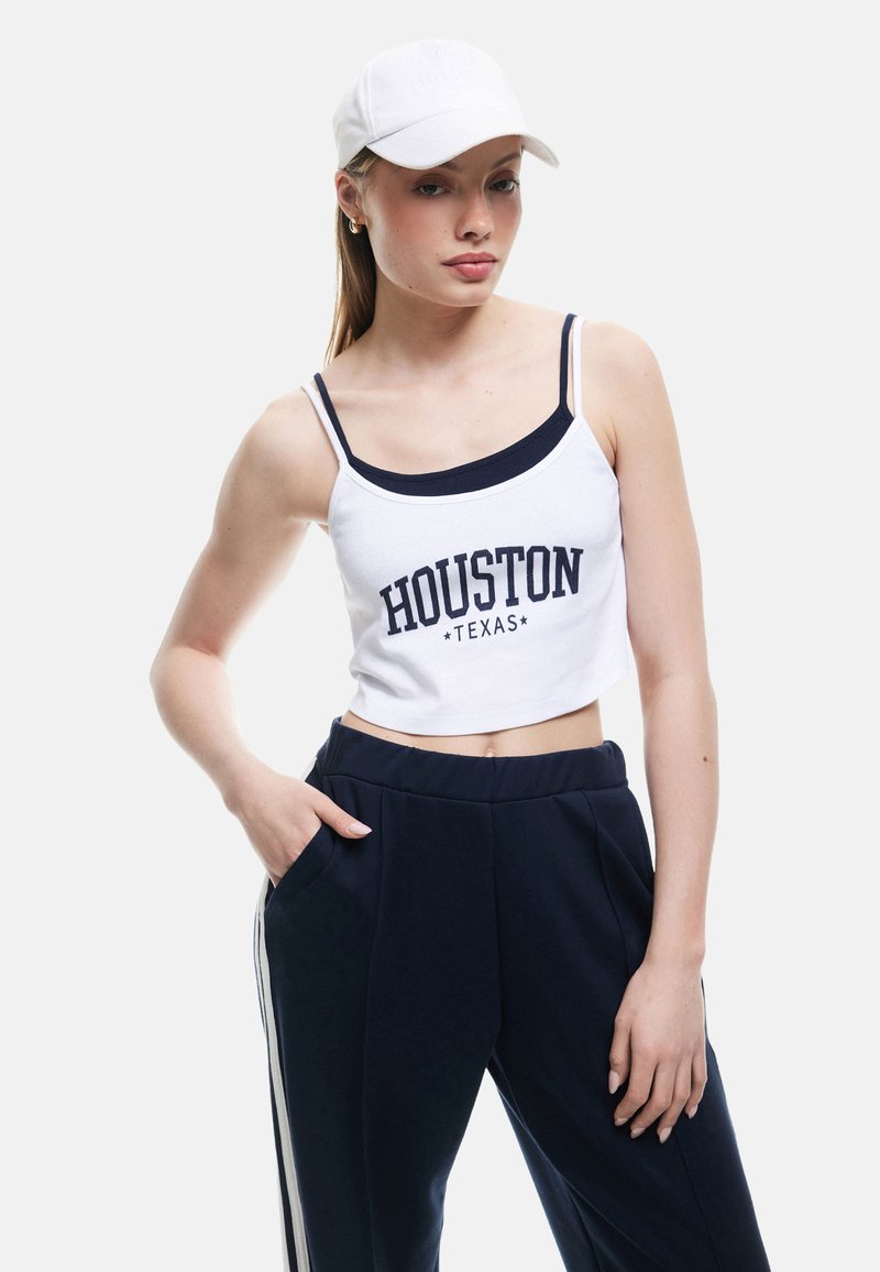 Biały krótki top na ramiączkach z napisem "HOUSTON TEXAS" w granatowym kolorze, zestawiony z granatowymi spodniami z białymi paskami po bokach. Modelka nosi białą czapkę.