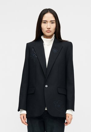 Vrouw met lang donker haar, gekleed in een zwarte blazer met subtiele sterren- en vogelborduursels, gedragen over een witte coltrui.