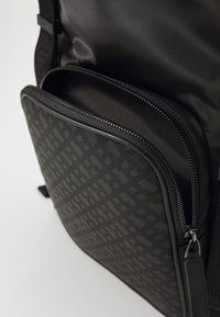Black backpack with a textured front pocket featuring a discernible pattern. Smooth material, zipper closures, and adjustable shoulder strap.