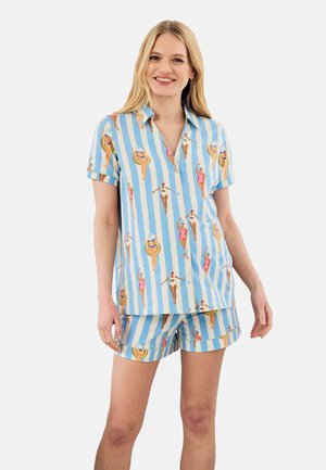 Kvinde iført blå- og hvide stribede kortærmede skjorte og shorts med svømmerillustrationer i vintage-stil, smilende mod en ensfarvet baggrund.