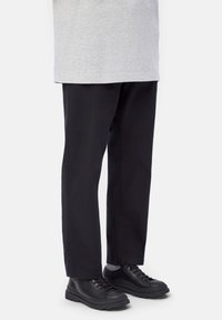 Persona che indossa pantaloni neri a gamba larga, calze grigie, scarpe in pelle nera con lacci e un top grigio chiaro su uno sfondo neutro.