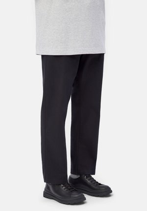 Person wearing black wide-leg pants, grey socks, black leather lace-up shoes, and a light grey top against a plain background.