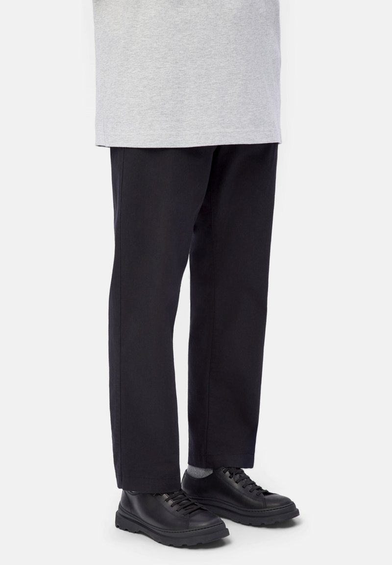 Personne portant un pantalon large noir, des chaussettes grises, des chaussures noires en cuir à lacets et un haut gris clair sur un fond uni.