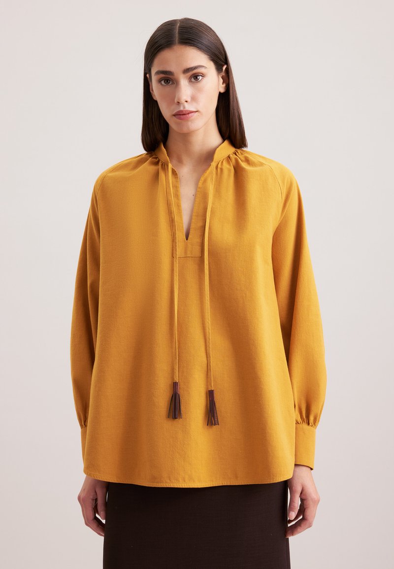 Senfgelbes Bluse mit lässiger Passform, langen Ärmeln, Stehkragen und vorderem Bindeband mit Quasten. Glatte Textur, minimales Design.
