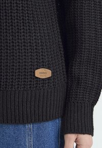 Czarny sweter dzianinowy z prążkowanymi mankietami i dołem. Na przodzie znajduje się brązowa skórzana naszywka z logo "OXMO". Luźny krój i teksturowany materiał.