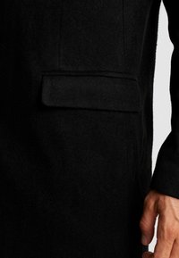 Cappotto in lana nera caratterizzato da una texture liscia, vestibilità aderente e una tasca applicata sul lato sinistro, con un dettaglio sartoriale sulle maniche.