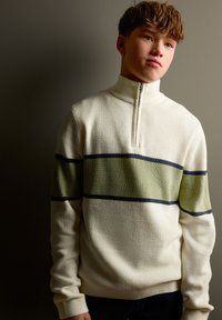 Pull blanc côtelé avec un col montant, arborant des rayures horizontales vert olive et bleu marine sur la poitrine. Texture douce et coupe ajustée.