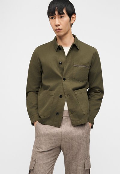 Άνδρας που φοράει πουκάμισο-ζακέτα σε χρώμα ελιάς με τσέπες και μπεζ παντελόνι τύπου cargo, στέκεται με τα χέρια στις τσέπες μπροστά σε λευκό φόντο.