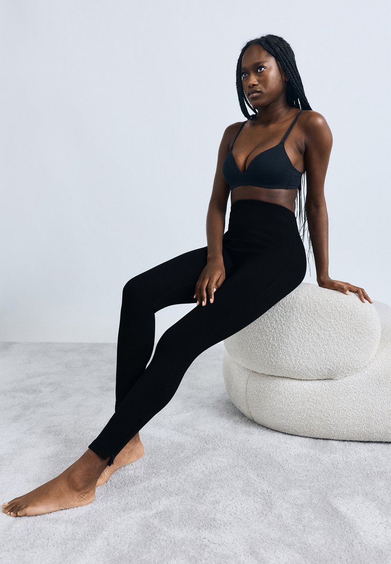 Sorte elastiske leggings med høj talje og tætsiddende design, parret med en matchende bralette, siddende på en tekstureret cremefarvet puf.