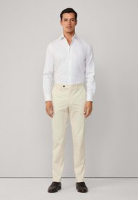 Un uomo sta in piedi di fronte, indossando una camicia bianca, pantaloni beige chiaro e scarpe marroni scure, su uno sfondo grigio chiaro uniforme.