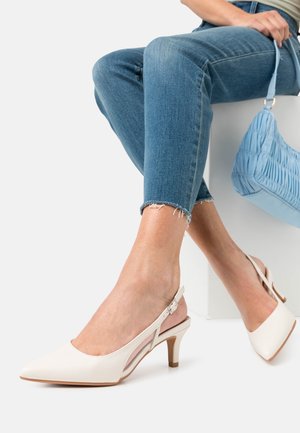 Femme portant des escarpins blancs à bouts pointus et talons bas, un jean bleu court, tenant un sac à main bleu clair froncé, assise.