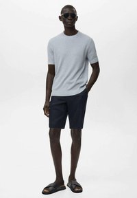 Maglione corto a maniche corte grigio chiaro a costine, pantaloni corti blu scuro e sandali neri; modella con occhiali da sole, in piedi con le mani in tasca.