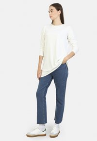 Weißes Langarmoberteil, schichtweise über blauen, geraden Jeans getragen, kombiniert mit weißen Sneakers mit grünen Akzenten und beigen Gummi-Sohlen.