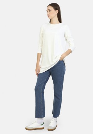White long-sleeve top layered over blue straight-leg jeans, worn with white sneakers featuring green accents and tan rubber soles.