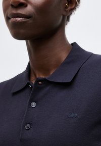 Κοντινό πλάνο ενός ατόμου που φοράει σκούρο ναυτικό μπλουζάκι πόλο με γιακά, δύο κουμπιά κλειστά και ένα μικρό κεντημένο λογότυπο στο στήθος.