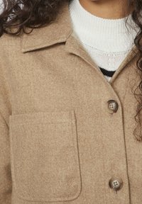 Giacca in misto lana color beige con colletto, chiusura frontale a bottoni e una tasca sul petto. Presenta una finitura morbida e testurizzata.