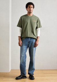 T-shirt verde de algodão por cima de uma camisa de mangas longas branca, combinado com jeans azul e sapatos pretos slip-on. Design simples, ajuste casual.